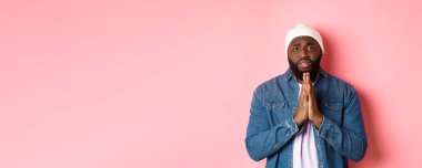 Üzgün, Afro-Amerikalı bir adam yardım istiyor, el ele tutuşup dua ediyor, üzgün bir şekilde kameraya bakıyor ve yalvarıyor, pembe arka planda duruyor..