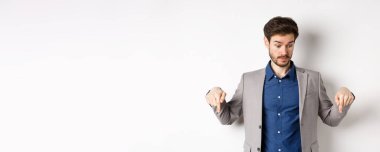 Şaşkın iş adamı yukarı kaş kaldırarak aşağı bakıyor ve tuhaf bir şey görüyor, takım elbiseli beyaz arka planda duruyor..