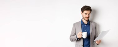 Takım elbiseli bir işadamı gazete okuyor ve kahve içiyor, kameraya güvenerek gülümsüyor, beyaz arkaplan.