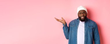 Sinirli ve kafası karışmış siyah adam tartışıyor, elini kaldırıyor ve kameraya şaşkın şaşkın bakıyor, ne olduğunu anlayamıyor, pembe arka planda duruyor..