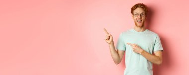 Kızıl saçlı neşeli bir gencin portresi, gözlük takıyor, sol üst köşeyi işaret ediyor ve gülümsüyor, pembe arka planda duruyor..