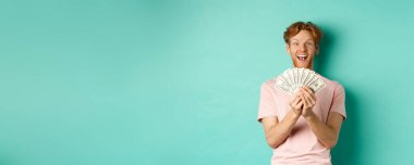 Heyecanlı genç adam ödül parası kazanıyor, nakit sayıyor ve dolarlara hayran bakıyor, turkuaz arka planda duruyor..