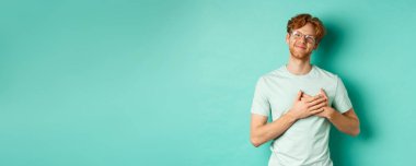 Kızıl saçlı ve gözlüklü yakışıklı genç adam, kalbe sarılmış ve gülümsüyor, teşekkür ediyor, minnettar ve duygulanmış hissediyor, turkuaz arka planda duruyor..