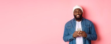 Yakışıklı Afro-Amerikalı adam akıllı telefon kullanıyor, kameraya gülümsüyor, pembe arka planda duruyor..