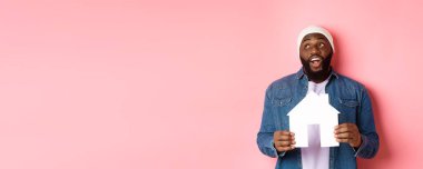 Emlak konsepti. Yakışıklı Afro-Amerikalı adam evini düşlüyor, ev modelini tutuyor ve sol üst köşeye bakıyor, resim dairesi, pembe arka plan..
