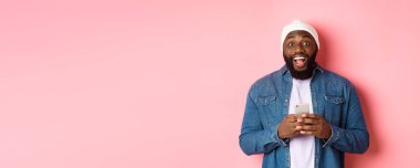 Teknoloji ve online alışveriş konsepti. Şaşkın genç siyahi adam cep telefonu kullanıyor, kameraya bakıyor. Mesajı okuduktan sonra şaşkın ve mutlu. Pembe arka plan..