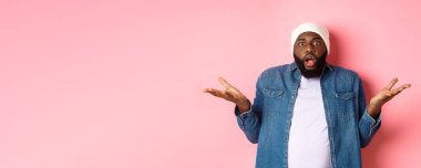 Kafası karışmış ve şok olmuş Afro-Amerikan erkeği ellerini yana açtı ve yere düştü, kameraya hayranlık ve şaşkınlıkla baktı, pembe arka planda hiçbir şey bilmeden durdu..