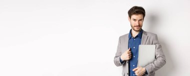 Başarılı bir iş adamı şık bir takım elbise içinde dizüstü bilgisayar taşıyor ve kameraya kendinden emin bakıyor, gülümsüyor memnun, beyaz bir altyapısı var..