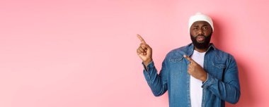 Şüpheci ve şüpheci siyah adam kameraya bakıyor emin değil, parmaklarını sol üst köşedeki belirsiz bir şeye doğrultuyor, pembe arka planda duruyor..
