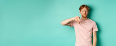 Hayal kırıklığına uğramış kızıl saçlı adam, baş parmağını kaldırmış, kaşlarını çatmış, şüpheci, hoşnutsuz ve hoşnutsuz görünüyor, turkuaz arka planda duruyor..