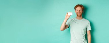 Alışveriş konsepti. Plastik kredi kartı gösteren, gülümseyen, turkuaz arka planda duran, tişörtlü yakışıklı kızıl adam..