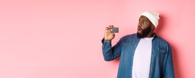 Alışveriş konsepti. Sakallı Afro-Amerikan bir adam kredi kartına bakarken şok olmuş, pembe arka planda duruyor..