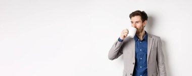 Takım elbiseli yakışıklı bir işadamı ofis kupasından kahve ya da çay içiyor, logoya bakıyor, beyaz arka planda duruyor..