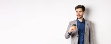 Takım elbiseli yakışıklı iş adamı kahve içiyor ve kameraya bakıyor, beyaz arka planda duruyor..