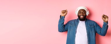 Mutlu genç Afro-Amerikalı adam kulaklıkla dans ediyor ve müzik dinliyor, yumruklarını sallıyor ve gülümsüyor, pembe arka planda duruyor..