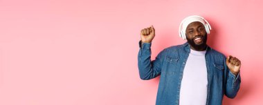 Mutlu siyah adam dans ediyor ve kulaklıkla müzik dinliyor. Neşeli gülümsüyor, sokak tarzı kıyafetlerle pembe arka planda duruyor..