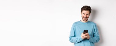Akıllı telefon kullanan, gülen, kameraya gülümseyen, beyaz arka planda duran yakışıklı bir adam..