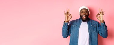 Bereli ve gömlekli etkileyici siyahi adam, tamam işaretleri gösteriyor ve kameraya şaşkın şaşkın bakıyor, gülümsüyor, harika bir teklif sunuyor, ürün öneriyor, pembe arka plan..