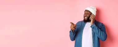 Cep telefonuyla konuşan yakışıklı, solu işaret eden ve gülümseyen, pembe arka planda duran siyahi adam..