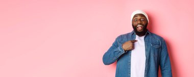 Hipster bereli şaşkın siyah adam kendini işaret ediyor, kameraya şaşkın şaşkın bakıyor, pembe arka planda duruyor..