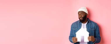 Emlak konsepti. Şüpheli siyah erkek model kağıt ev modelini tutuyor, şüpheli ve şüpheli görünüyor, daireyi arıyor, pembe arka planda duruyor..