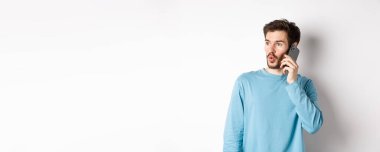 Teknoloji konsepti. İlgi çekici genç adam soldan etkilenmiş görünüyor, cep telefonuyla konuşuyor, birini arıyor, beyaz arka planda duruyor..