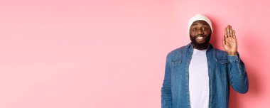 Dostça gülümseyen siyah adam merhaba diyor, el sallıyor, seni selamlıyor, pembe arka planda duruyor..