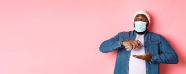 Covid-19, yaşam tarzı ve tecrit konsepti. Gülümseyen Afro-Amerikan maskeli adam dezenfektanla ellerini temizliyor, antiseptik kullanıyor ve kameraya bakıyor, pembe arka plan.