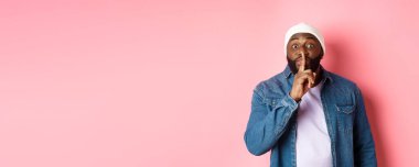 Bereli ve kot gömlekli yakışıklı genç adam sürprizi hazırla, sessiz ol ve kameraya bak, sessiz ol, pembe arka plan.