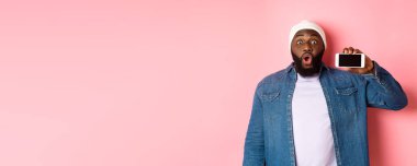 Çevrimiçi alışveriş ve teknoloji kavramı. Şaşırmış siyah adam telefon ekranını yatay olarak gösteriyor ve kameraya bakıyor heyecanlı, pembe arka planda duruyor..