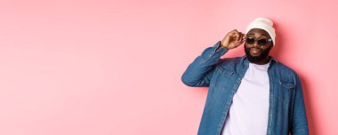 Havalı ve küstah Afro-Amerikan sakallı adam, kendinden emin görünüyor, güneş gözlüklerine dokunuyor ve pembe arka planda duran kameraya bakıyor lütfen..