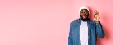 Dostça gülümseyen siyah adam merhaba diyor, el sallıyor, seni selamlıyor, pembe arka planda duruyor..