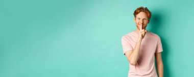 Kızıl saçlı ve sakallı gülümseyen genç bir adam bir sırrı paylaşıyor, tabu jesti yapıyor ve sırıtıyor, sessiz olman için seni susturuyor, turkuaz arka planda dikiliyor..