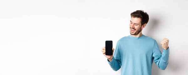 Электронная коммерция и концепция покупок. Счастливчик выигрывает онлайн, показывая пустой экран смартфона и триумф, говоря "да" с помощью кулачного насоса, стоя на белом фоне.