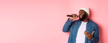 Bereli ve kot gömlekli yakışıklı siyah adam karaoke söylüyor, mikrofon tutuyor, pembe arka planda duruyor..
