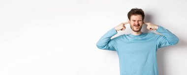 Sinirli genç adam korkunç seslerden kulaklarını tıkar, rahatsız edici gürültüden rahatsız olur, hoşnutsuz olur, beyaz arka planda durur..