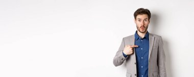 Kim ben mi? Takım elbiseli şaşırmış bir adam kendini inançsızlıkla işaret ediyor, kameraya bakıyor, beyaz arka planda duruyor..