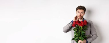Takım elbiseli genç adam elinde bir buket çiçekle Sevgililer Günü 'nde randevuyu bekliyor, beyaz arka planda dikiliyor..