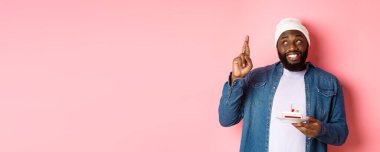 Umut dolu siyahi adam doğum gününü kutluyor, parmaklarını çaprazlayarak dilek tutuyor, elinde mumla doğum günü pastası tutuyor, pembe arka planda duruyor..