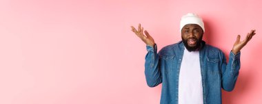 Kafası karışmış ve rahatsız olmuş siyah adam şikayet ediyor, omuz silkiyor ve ellerini kaldırıyor, hayal kırıklığıyla bakıyor, pembe arka planda duruyor..