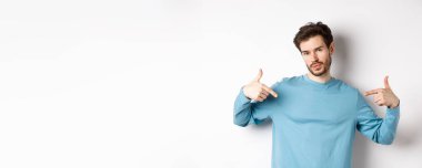 Sakallı genç adam kendine güveniyor, kendini gösteriyor ve kendini beğenmiş bir ifade takınıyor, beyaz arka planda havalı ve küstah duruyor..