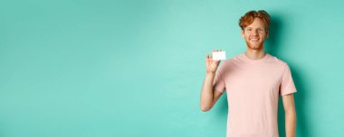 Yakışıklı genç adam gülümsüyor ve plastik kredi kartı gösteriyor. Tişörtünün içinde turkuaz arka planda duruyor..