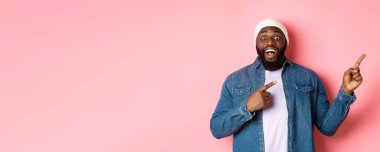 Heyecanlı ve şaşırmış siyah adam harika bir teklif sunuyor, parmaklarını fotokopi alanına doğrultuyor, pembe arka planda hippi beresi ve kot gömlek giyiyor..