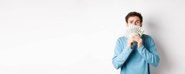 Endişeli genç adam yalvaran yüz ifadesiyle, para gösteriyor, beyaz arka planda mavi kazakla dikiliyor..