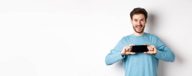 E-ticaret ve alışveriş konsepti. Gülümseyen neşeli adam yatay pozisyonda boş akıllı telefon ekranı gösteriyor, kameraya mutlu bakıyor, beyaz arka plan.