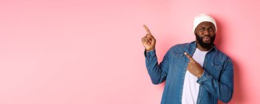 Kafası karışmış ve hayal kırıklığına uğramış Afro-Amerikan bir adam yüzünü ekşitmiş, sol üst köşedeki afişi işaret ediyor, iğrenç yüzüyle reklamını yapıyor, pembe arka planda duruyor..