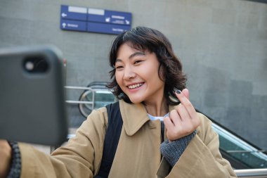 Portrait of stylish korean girl, student takes selfie on street, holds smartphone and poses on its camera, makes video on tourist trip, smiles with joy.