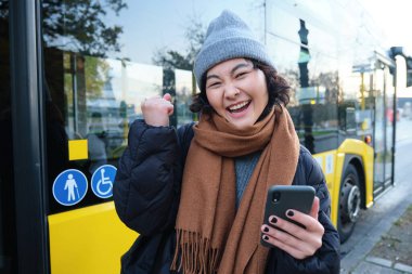 Enthusiastic girl rejoice, reads message on mobile phone and celebrates, stands near her bus on public transport stop and looks excited, posing in warm winter clothes.