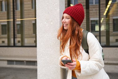 Stylish urban girl using mobile phone app, standing in city, waiting for taxi, looking at smartphone application, texting message.