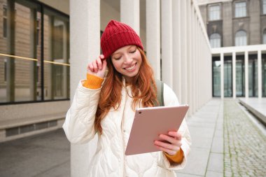 Happy redhead girl in red hat, walks around city with digital tablet, connects to public internet wifi and looks for route, looks at map on her gadget.